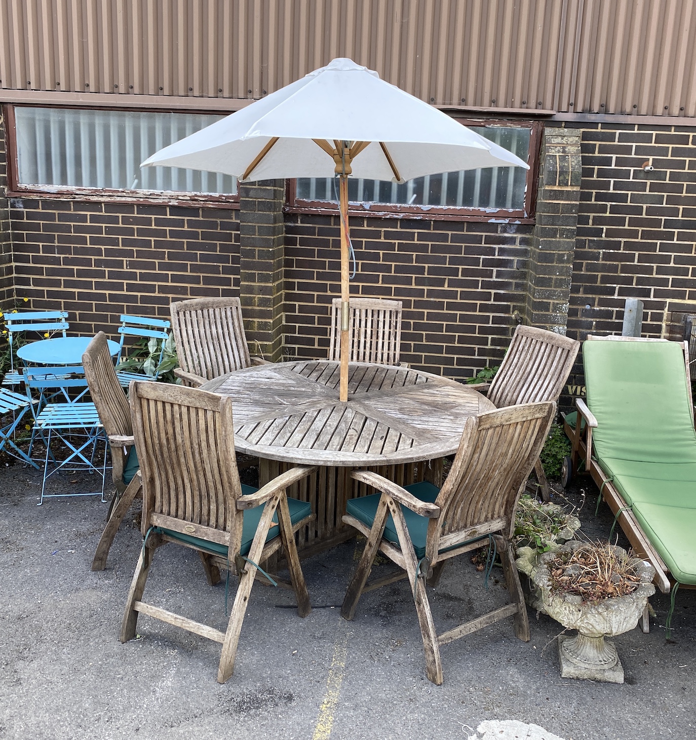 A weathered teak circular garden table, diameter 153cm, height 74cm together with six folding elbow chairs with cushions, stamped 'Regency' and a parasol